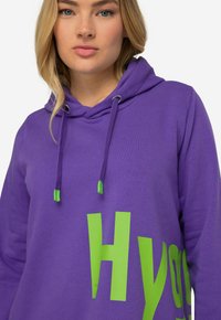 Paarse hoodie gemaakt van zachte stof, met een voorzak, verstelbare groene trekkoorden en grote groene tekstgrafiek op de voorkant.