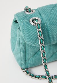 Turquoise denim handbag with a quilted design, silver hardware, and a chain strap featuring turquoise accents. Textured surface detail visible.