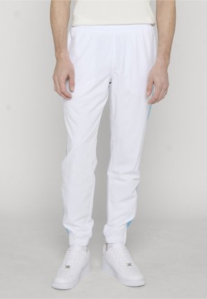 Osoba ubrana w białe spodnie dresowe z elastycznymi mankietami i białe trampki, stojąca na jasnoszarym tle.