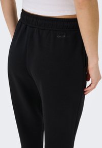 Κοντινό πλάνο ενός ατόμου που φορά μαύρο ελαστικό παντελόνι φόρμας με λευκό ριμπ κορμάκι, εμφανίζοντας το δεξί χέρι στο πλάι.