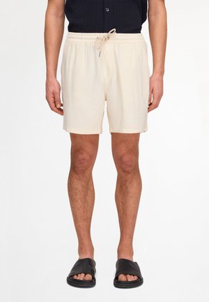 Mężczyzna ubrany w beżowe szorty z troczkiem, czarną koszulę zapinaną na guziki i czarne klapki, stojący na białym tle.