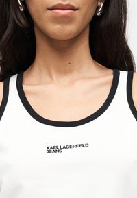 Κοντινή λήψη μιας γυναίκας που φοράει λευκή φανέλα με μαύρο τελείωμα και την επιγραφή «KARL LAGERFELD JEANS» κεντημένη στο στήθος.