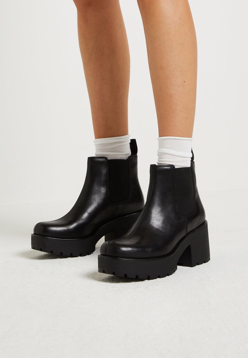 Jambes portant des chaussettes blanches et des bottines noires à talons épais avec des côtés élastiques sur un sol et un fond clairs.