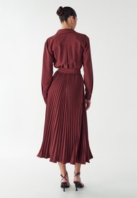 Bordeaux väriset lyhyet hameet, joissa on kauluspaitakupu, vyötärölle sidottu vyö ja laskeutuva hame. Sujuva kangas ja kerrokselliset laskokset korostavat sen siluettia.