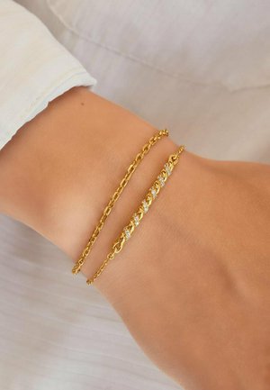 Handgelenk mit zwei zarten goldenen Gliederarmbändern, eines davon mit kleinen eingearbeiteten Kristallen, vor hellem Stoffhintergrund.