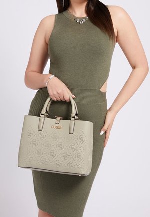 Frau in olivgrünem ärmellosem Kleid hält eine hellbeige Guess-Handtasche mit diamant- und floralen Ausschnittmustern.