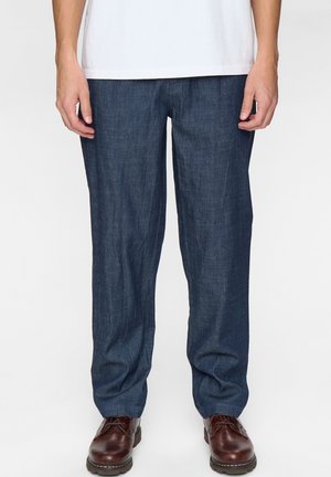 Uomo con pantaloni larghi blu scuro, camicia bianca e scarpe di pelle marrone scuro, in piedi davanti a uno sfondo bianco.