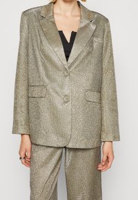 Blazer metálico dorado con tela texturizada, cierre de un solo botón, solapas muesca y dos bolsillos frontales. Combinado con pantalones a juego.