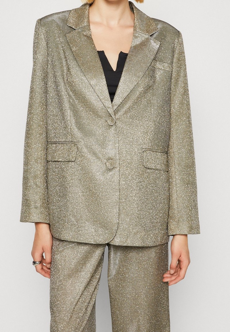 Blazer metálico dorado con tela texturizada, cierre de un solo botón, solapas muesca y dos bolsillos frontales. Combinado con pantalones a juego.