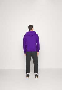 Sudadera con capucha morada, con un bolsillo tipo canguro, puños y dobladillo acanalados. Combinada con pantalones oscuros y zapatillas, sobre un fondo liso.