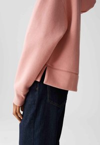 Vista laterale di una persona che indossa un top lungo rosa con orlo a spacco e jeans blu scuro, con la mano rilassata lungo il corpo.