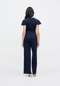 Mono azul marino con mangas cortas estilo volante, cintura entallada y pantalones de pierna ancha. Textura lisa con cierre de cremallera en la parte trasera.