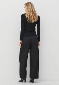 Ženska s svetlim lasmi, oblečena v črno dolgosleečno majico, široke črne hlače in črne čevlje s peto, obrnjena proti beli steni, vidna je njena hrbtna stran.