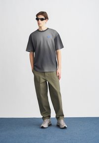 Γκρι μπλουζάκι με διαβάθμιση, άνετη γραμμή, κοντά μανίκια, με μικρό μπλε λογότυπο. Ελαιδί παντελόνια, ίσια γραμμή, και γκρι αθλητικά παπούτσια.