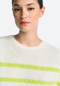 Pulover bej deschis, realizat din material moale și pufos, cu trei dungi orizontale îndrăznețe de culoare verde lime. Guler rotund și margini reiate.