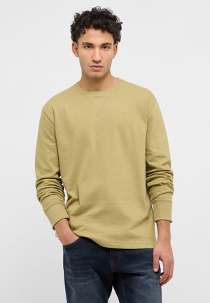 Mann mit lockigem Haar, der einen hellolivgrünen Pullover und dunkelblaue Jeans trägt, steht mit einer Hand in der Tasche vor einfarbigem Hintergrund.