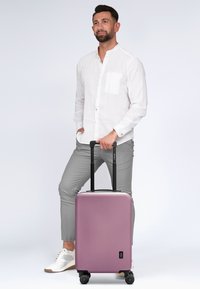 Hartschalenkoffer in mattem Lavendel-Finish mit schwarzem ausziehbarem Griff und Rädern, mit einem kleinen Logodetail auf der Vorderseite.