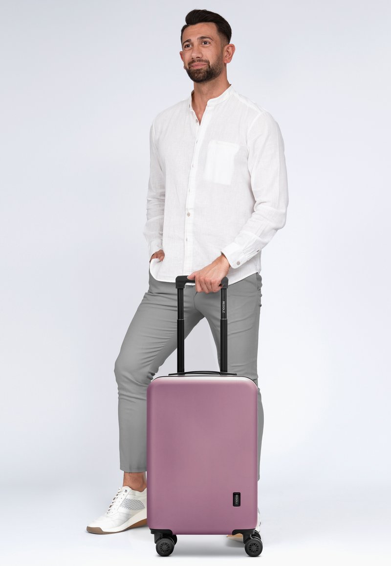 Hartschalenkoffer in mattem Lavendel-Finish mit schwarzem ausziehbarem Griff und Rädern, mit einem kleinen Logodetail auf der Vorderseite.