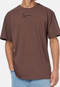 Brązowa bawełniana koszulka z krótkim rękawem, z czarnym haftowanym logo na piersi i luźnym krojem. Dół jest nieco dłuższy z tyłu.