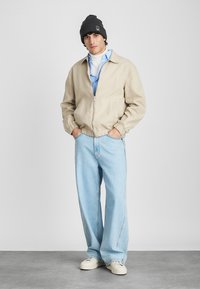 Beige bomber jacket with elastic cuffs, light blue button-up shirt, oversized light blue jeans, black beanie, and white sneakers.