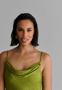 Grøn satin camisole med cowl-udskæring og tynde stropper, der har et subtilt slangemønster og en glat tekstur. Model med kort hår.