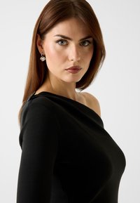 Vestito nero con spalle scoperte, caratterizzato da una silhouette aderente, texture liscia e dettagli decorativi sulla spalla. Modella con capelli castani lunghi.