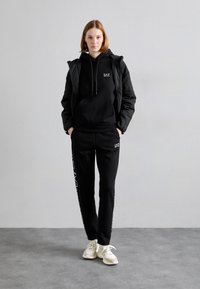 Melnas hoody un sporta bikses ar sportisku dizainu; satur logo un kontrastējošas baltas akcentus, saskaņotas ar gaišām kedām.