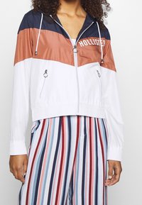 Färgad vindtäta jacka i marinblått, rostfärgat och vitt med dragkedja fram, elastiska manschetter och två sidofickor. Funktioner "HOLLISTER"-logotyp i vitt.