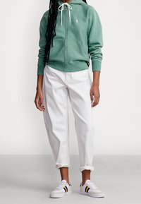 Felpa verde con zip e lacci bianchi, abbinata a pantaloni di jeans bianchi arrotolati. Le scarpe sono sneakers bianche con strisce gialle e navy.