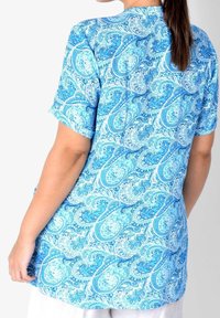 Camicia a maniche corte con stampa paisley blu, caratterizzata da un design fluido. Il tessuto ha una consistenza liscia e una palette di colori vivaci di blu.
