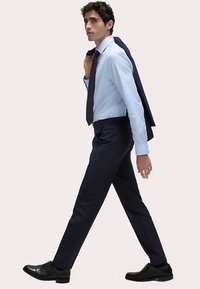 Skræddersyede marineblå bukser, lyseblå skjorte, mørkt slips og sorte festlige sko. Modellen bærer en marineblå blazer over skulderen og går i profil.