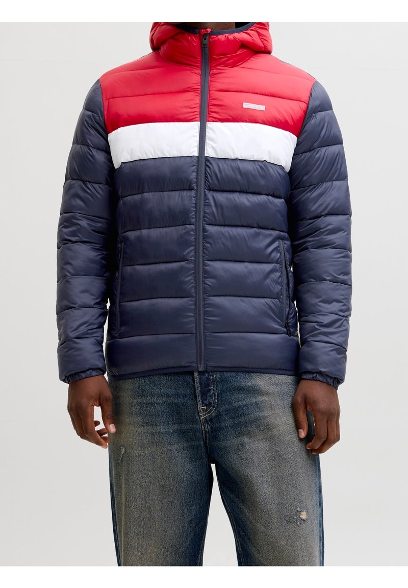 Homme portant une veste matelassée bleu marine, blanche et rouge avec une capuche et une fermeture éclair à l'avant, associée à un jean bleu délavé.