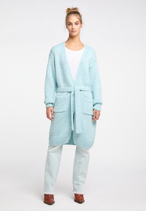 Vrouw draagt lichtblauwe gebreide cardigan met ceintuur, wit overhemd, lichtgewassen spijkerbroek en bruine laarzen, staand tegen een witte achtergrond.