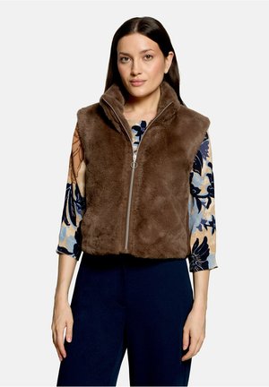 Frau trägt eine braune, flauschige Reißverschlussweste über einem floralen Langarmshirt und dunklen Hosen, steht mit neutralem Gesichtsausdruck.