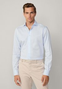 Camicia azzurra chiara a maniche lunghe in tessuto morbido, con colletto a punta e chiusura a bottoni, abbinata a pantaloni beige.