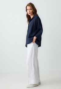 Chemise en lin bleu marine avec col et poches latérales, associée à un pantalon large en lin blanc et des baskets en toile beige. Design minimaliste.