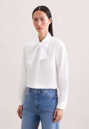 Kobieta ubrana w białą bluzkę z długim rękawem z dużą kokardą przy kołnierzyku i niebieskie jeansy z wysokim stanem, stojąca na jednolitym tle.