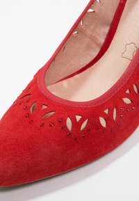 Chaussure plate en daim rouge à bout pointu, motifs décoratifs découpés en forme de feuilles et petits strass rouges le long du bord supérieur.