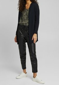 Long black cardigan over a green patterned top, paired with black faux leather pants and white sneakers. Soft textures, relaxed fit.