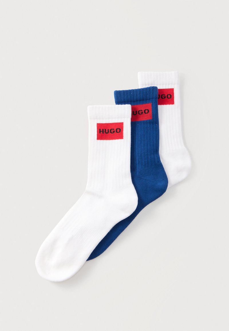 Trois chaussettes montantes côtelées, deux blanches et une bleue, chacune avec un patch logo rouge rectangulaire portant l'inscription « HUGO » près du haut du revers.