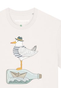 Witte t-shirt met een meeuw in een gestreept shirt en groene hoed, staande op een fles met een papieren boot en golfontwerp.