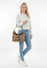 Woman in light gray sweater and blue jeans holding a large tan leather shoulder bag, smiling and standing with crossed legs.