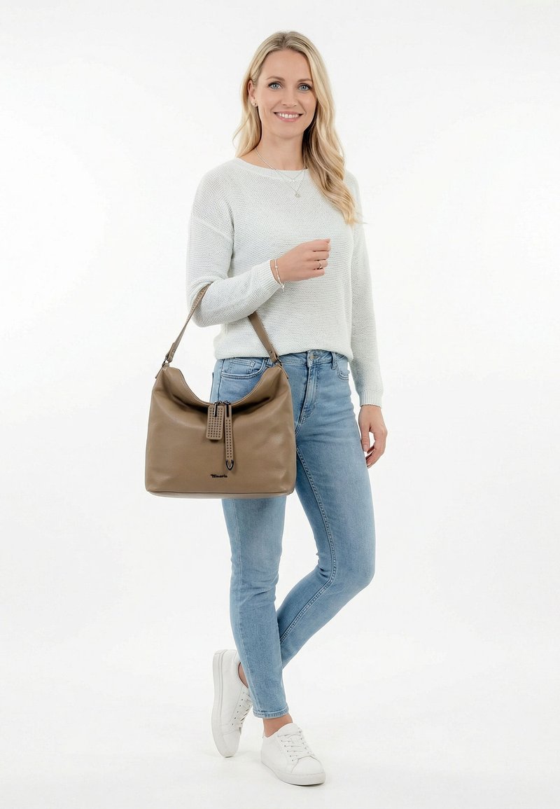 Woman in light gray sweater and blue jeans holding a large tan leather shoulder bag, smiling and standing with crossed legs.