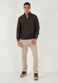 Pullover zip-up marrone scuro con texture a costine, abbinato a pantaloni beige chiari e scarpe da ginnastica multicolori con lacci neri.