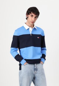 Polo-skjorte med horisontale striper i lyseblå og marine, med hvit krage og liten brodert logo på brystet. Laget av bomull.