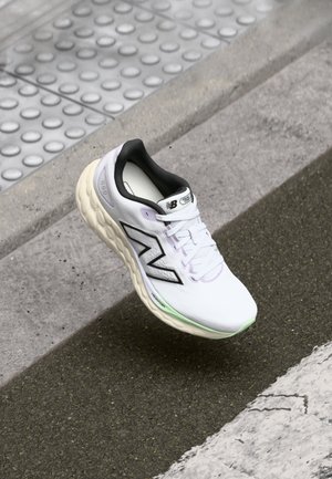 Biały but do biegania New Balance z czarnym logo i amortyzowaną podeszwą na miejskim chodniku blisko płytek dla niewidomych i krawężnika.
