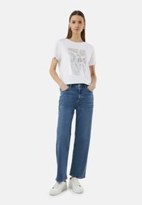 White T-shirt with a silver graphic design, paired with blue wide-leg jeans. The model wears white sneakers and has a neutral background.