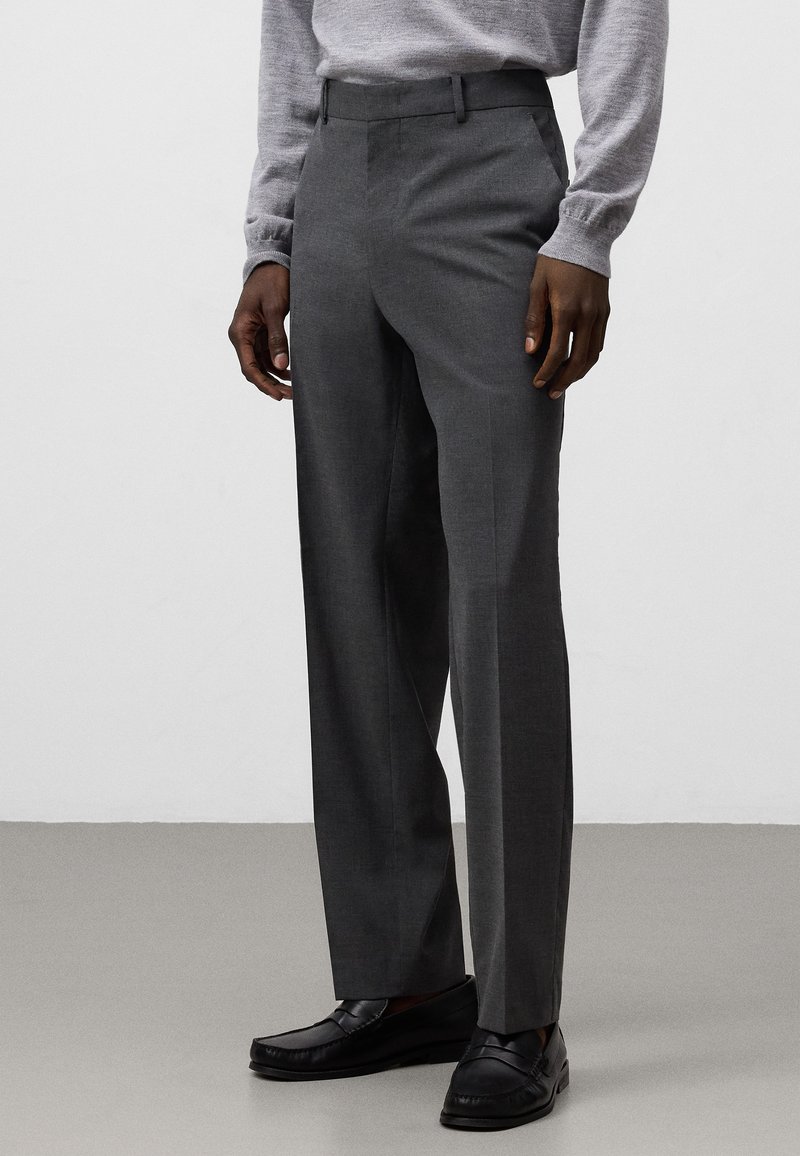 Pantaloni grigi su misura con un design a gamba dritta, realizzati in tessuto strutturato. Presentano passanti per cintura e tasche laterali, abbinati a mocassini neri.