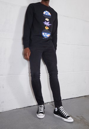 Persona vestida con sudadera negra con diseño de globo y flor en azul y naranja, vaqueros pitillo negros y zapatillas altas negras.