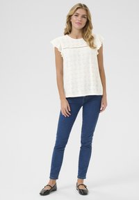 T-shirt blanc en eyelet avec des manches courtes à volants, col rond et motif floral perforé. Associé à un jean skinny bleu foncé et des ballerines noires.